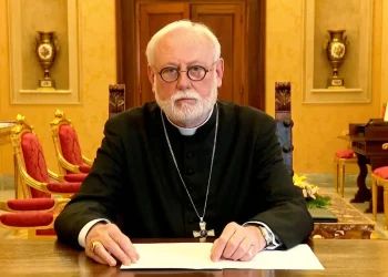 Arcivescovo Gallagher al tavolo, indossa abito clericale e occhiali.