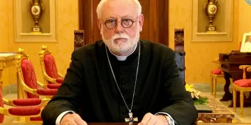 Arcivescovo Gallagher al tavolo, indossa abito clericale e occhiali.