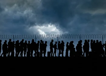 Silhouette di persone, migranti, in fila davanti a un recinto spinato