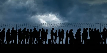 Silhouette di persone, migranti, in fila davanti a un recinto spinato