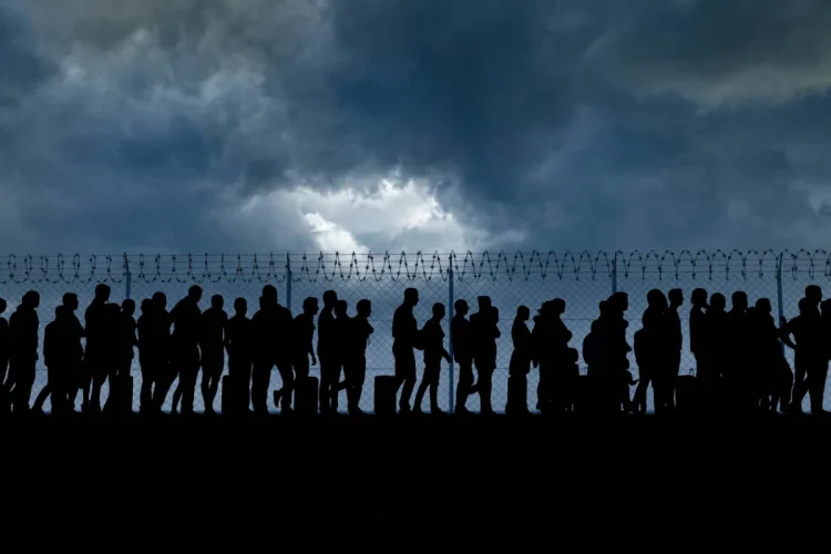 Silhouette di persone, migranti, in fila davanti a un recinto spinato