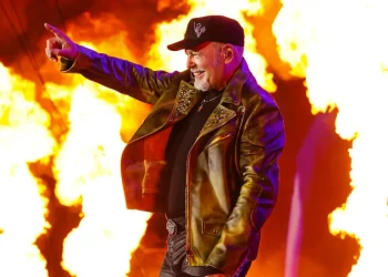 Vasco Rossi sorride su palco con fiamme, indica il pubblico.