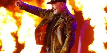 Vasco Rossi sorride su palco con fiamme, indica il pubblico.