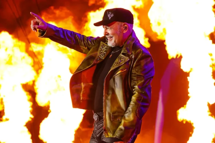 Vasco Rossi sorride su palco con fiamme, indica il pubblico.