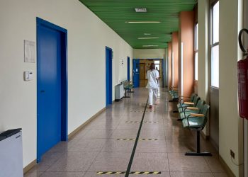 A healthcare worker walking down an empty hospital corridor with blue doors and green ceiling.