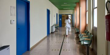 A healthcare worker walking down an empty hospital corridor with blue doors and green ceiling.