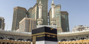 A large group of pilgrims gather at the Kaaba in Mecca, Saudi Arabia, during the Hajj pilgrimage under a clear blue sky.