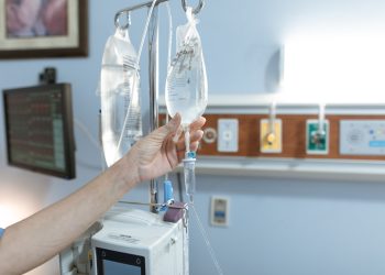 Close-up image of an IV drip and hand in a hospital room, illustrating medical care.