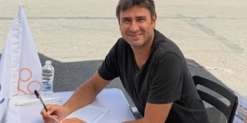 Alessandro Di Battista sorride seduto ad un tavolo all'aperto, firma documenti.