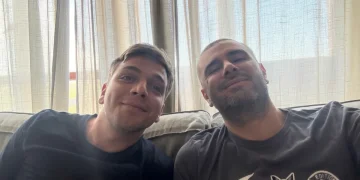 Due uomini sorridenti seduti sul divano in casa, atmosfera serena. Di Battista: "Sto molto meglio, finalmente sono a casa"