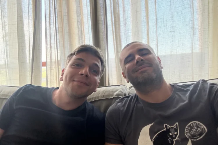 Due uomini sorridenti seduti sul divano in casa, atmosfera serena. Di Battista: "Sto molto meglio, finalmente sono a casa"