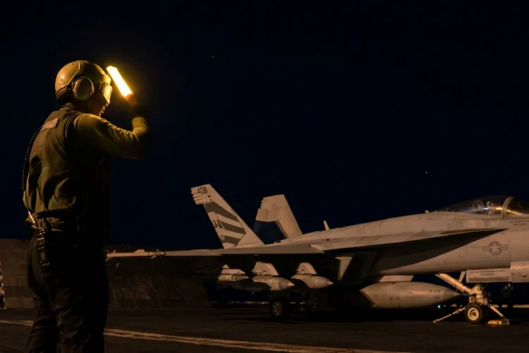 Membro personale di terra dirige F-18 Hornet su portaerei di notte.