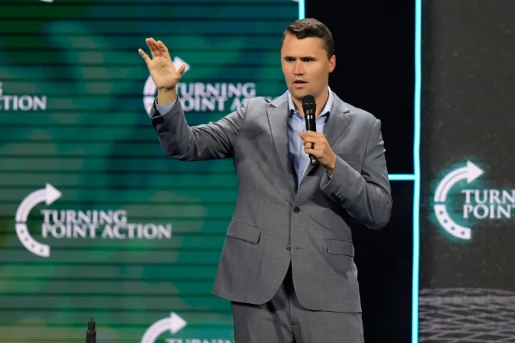 Charlie Kirk parla al microfono in completo grigio su un palco.