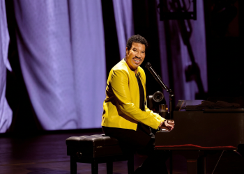 Lionel Richie suona il pianoforte sorridendo sul palco con giacca gialla. Lionel Richie ricoverato in terapia intensiva a St. Louis dopo il concerto