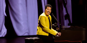 Lionel Richie suona il pianoforte sorridendo sul palco con giacca gialla. Lionel Richie ricoverato in terapia intensiva a St. Louis dopo il concerto