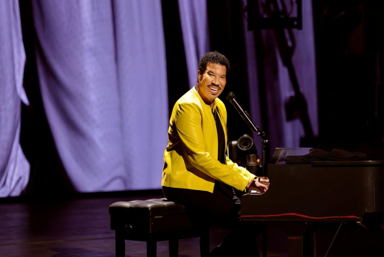 Lionel Richie suona il pianoforte sorridendo sul palco con giacca gialla. Lionel Richie ricoverato in terapia intensiva a St. Louis dopo il concerto