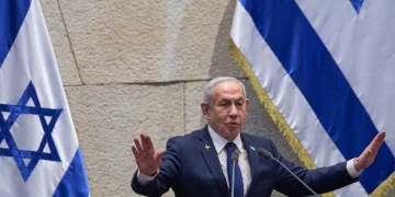 Benjamin Netanyahu parla con gesti energici, bandiere di Israele dietro.