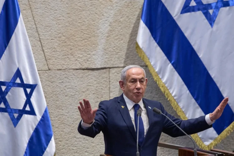 Benjamin Netanyahu parla con gesti energici, bandiere di Israele dietro.