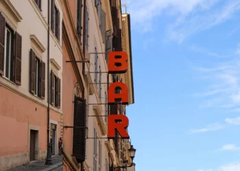 Insegna verticale rossa "BAR" su edificio romano antico, cielo azzurro
