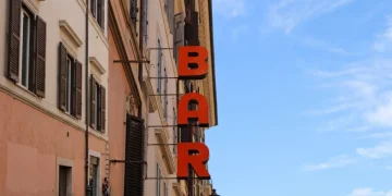 Insegna verticale rossa "BAR" su edificio romano antico, cielo azzurro