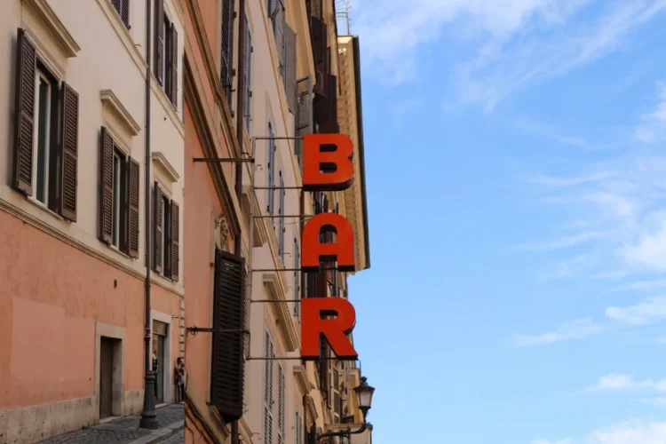 Insegna verticale rossa "BAR" su edificio romano antico, cielo azzurro