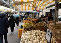 Colorful market stall in Italy featuring fresh potatoes, garlic, and fruit under striped awnings.