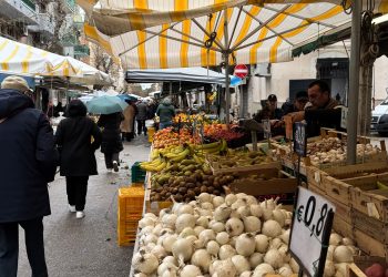 Colorful market stall in Italy featuring fresh potatoes, garlic, and fruit under striped awnings.