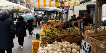 Colorful market stall in Italy featuring fresh potatoes, garlic, and fruit under striped awnings.