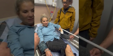 Donna ferita su barella intervistata con microfono in ospedale, uomo vicino.