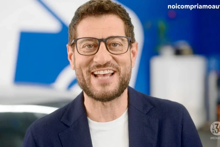 Enrico Papi sorride indossando occhiali da vista e giacca scura