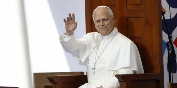 Papa Leone XIV saluta il pubblico, vestito di bianco, seduto.