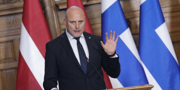 Kariņš, Primo Ministro lettone, parla al podio con bandiere europee.Lettonia, Lista Unita conquista 42 seggi e segna il miglior risultato nelle elezioni per un partito dal 1991